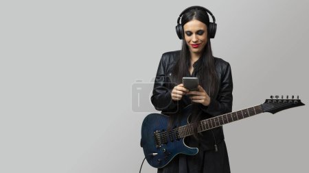 Téléchargez les photos : Une jeune femme, guitariste, aime écouter de la musique avec le casque sans fil et le téléphone portable qu'elle tient dans ses mains - en image libre de droit