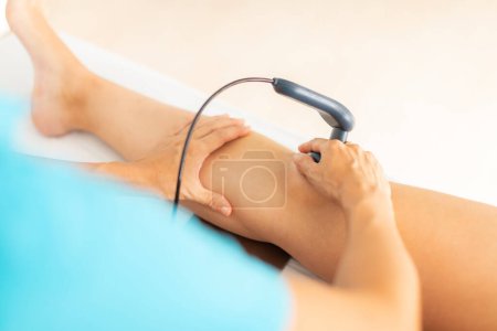 Téléchargez les photos : Un physiothérapeute applique des ondes radiofréquences avec une machine à diathermie pour soulager la douleur musculaire au genou de sa patiente - en image libre de droit