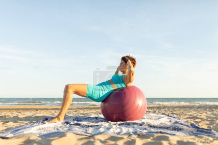 Photo for A woman performs sit-ups abs lying on a Pilates ball on the beach - Royalty Free Image