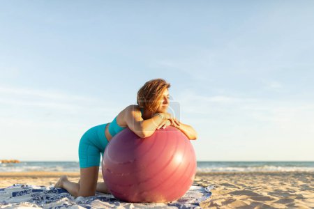 Foto de Una mujer descansa y se relaja durante una sesión de Pilates en la playa, apoyándose en una pelota de Pilates - Imagen libre de derechos