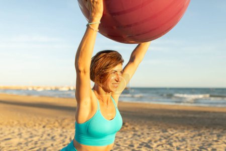 A woman performs stretching and balance exercises with a Pilates ball during a session on the beach
