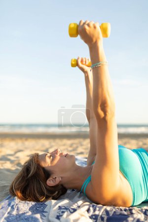 Photo for A woman performs stretching and relaxation exercises with light 1kg dumbell weights, lying on the beach - Royalty Free Image