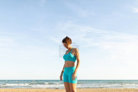 A woman rests after performing Pilates exercises on the beach, facing the sea