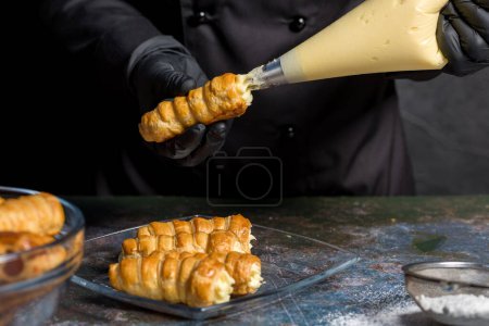 Téléchargez les photos : Le chef pâtissier remplit les tubes de pâte feuilletée avec de la crème à partir d'un sac à pâtisserie - en image libre de droit