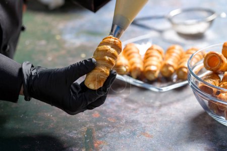 Téléchargez les photos : Le chef pâtissier remplit les tubes de pâte feuilletée avec de la crème à partir d'un sac à pâtisserie - en image libre de droit