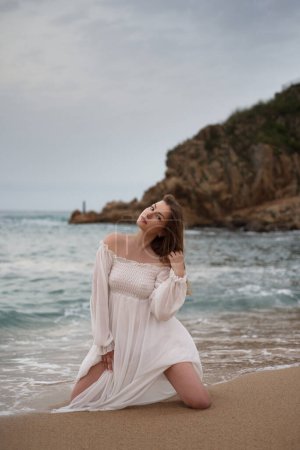 Téléchargez les photos : Une belle fille en robe blanche s'agenouille dans la mer sur fond de vagues. Temps couvert. Portrait vertical complet. Lumière du jour - en image libre de droit