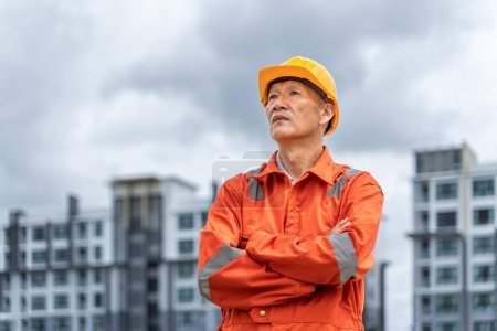 Téléchargez les photos : Ingénieur asiatique sérieux et déterminé d'âge moyen en combinaison orange avec un casque traversant ses bras tout en se tenant fièrement devant ses bâtiments à plusieurs étages avec un ciel nuageux en arrière-plan - en image libre de droit