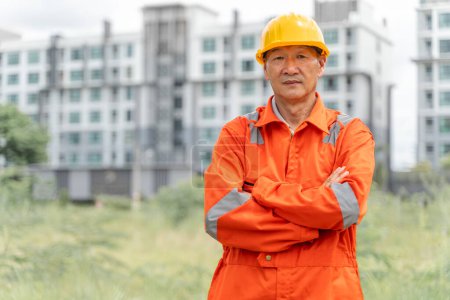 Téléchargez les photos : Ingénieur asiatique sérieux et déterminé d'âge moyen en combinaison orange avec un casque croisant les bras tout en se tenant fièrement devant ses bâtiments à plusieurs étages en arrière-plan - en image libre de droit