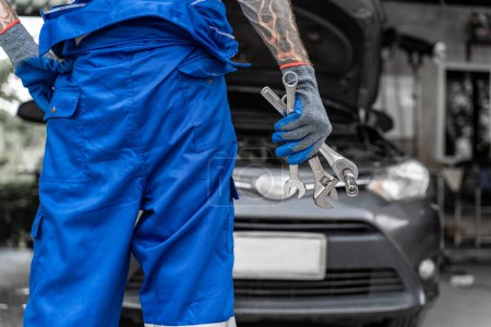 Close up hands and arms with tattoo of a male car mechanic from behind holding wranches with car in the background