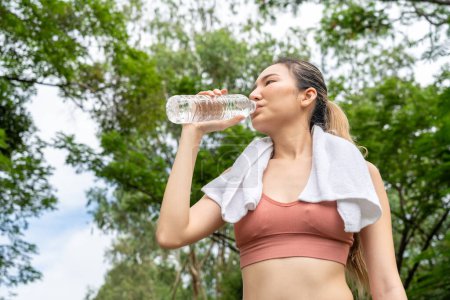Photo for Young Asian woman in fitness clothes stops to drink water from her plastic water bottle during her morning exercise run at a running track of a local park - Royalty Free Image
