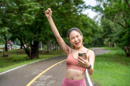 Téléchargez les photos : Heureuse jeune femme asiatique attrayante vérifiant le résultat de sa course matinale réussie et levant le bras droit pour célébrer sur une piste de course d'un parc local - en image libre de droit