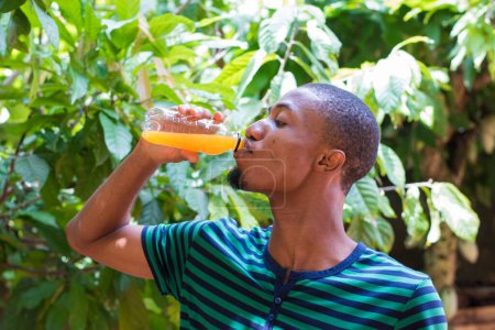 Foto de A handsome fair complexion African Nigerian male man drinking an orange juice from a transparent bottle with eyes closed from enjoying the refreshing drink - Imagen libre de derechos