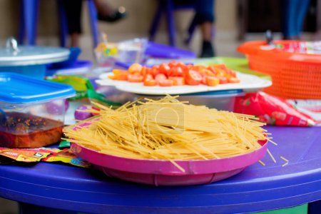 Téléchargez les photos : Un gros plan de nouilles spaghetti dans une assiette en plastique, placées sur une table de cuisine aux côtés d'autres ingrédients alimentaires pour préparer un repas sain, nutritionnel et délicieux - en image libre de droit