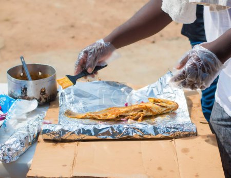 Téléchargez les photos : Mains d'un chef africain nigérian portant des gants en nylon transparent, préparant du poisson grillé avec un ingrédient pertinent tandis que les oignons et le gingembre font partie des épices - en image libre de droit