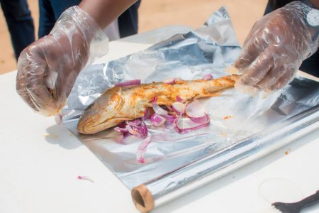 Téléchargez les photos : Mains d'un chef africain nigérian avec des gants en nylon transparent, la préparation de poissons grillés avec l'ingrédient pertinent tandis que les oignons et le gingembre font partie des épices - en image libre de droit