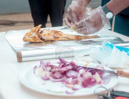 Téléchargez les photos : Mains d'un chef africain avec des gants en nylon transparent, préparer du poisson grillé avec un ingrédient pertinent avec des oignons et du gingembre dans le cadre des épices - en image libre de droit