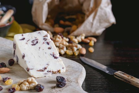 Wensleydale cheese with cranberries, red wine, honey, nuts, raisins on wooden cutting board. Black concrete background. Selective focus.