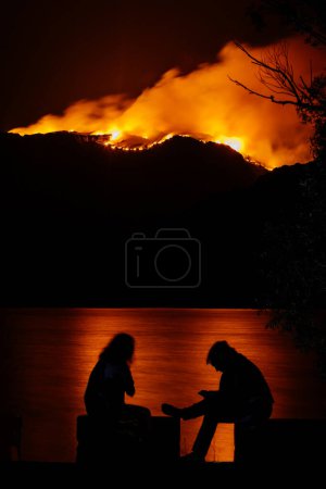 Silhouette zweier Frauen beim Beobachten von Waldbränden. Nächtliches Feuer im Wald mit Spiegelung im See.