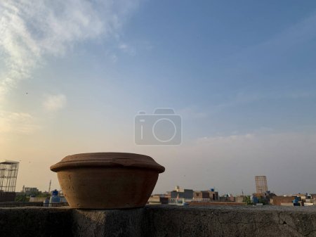 Mud water pot placed on the top of roof wall. Bird water feeder utensils with sunlight and a cloudy sky in the background.