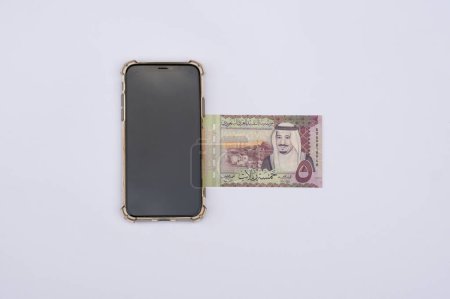 Saudi Arabia Riyal bills or bank notes placed under mobile phone. Isolated on a white background. Arabian country paper currency.