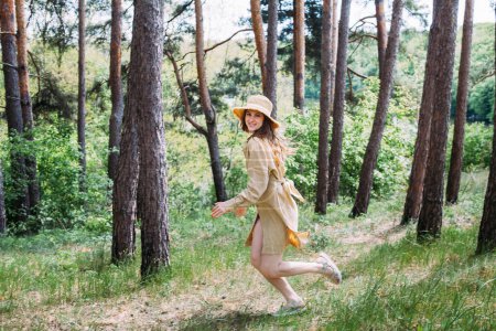 Foto de Una hermosa chica con el pelo rubio rizado en un vestido beige y sombrero corre por el bosque y sonríe. - Imagen libre de derechos