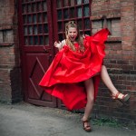 A cute young laughing girl with blond flowing long hair and beautiful make-up, in a long red dress, jumps along the street of the old city.