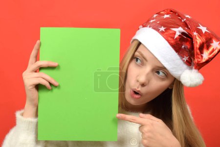Téléchargez les photos : Jeune femme en santa chapeau avec feuille verte de papier sur fond rouge - en image libre de droit