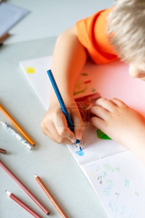 A child in an orange T-shirt draws with colored pencils on a white sheet.