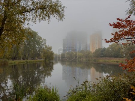 Téléchargez les photos : Tournage cinématographique d'une journée d'automne brumeuse dans un parc de la ville. Un lac au milieu du parc près des immeubles de grande hauteur et des arbres décorés de couleurs automnales et recouverts de brouillard. Photo de haute qualité - en image libre de droit