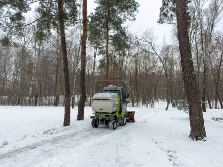 Téléchargez les photos : Les employés municipaux nettoient un parc municipal sur un tracteur. Service municipal de nettoyage de la neige, un petit tracteur mignon avec une brosse rotative nettoie une route dans le parc de la ville de la neige fraîche tombée le jour d'hiver. Haut - en image libre de droit