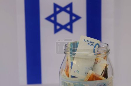 Téléchargez les photos : Pot en verre avec shekel israélien sur le fond du drapeau d'Israël. Concept : épargne personnelle, système bancaire, inflation, monnaie nationale - en image libre de droit
