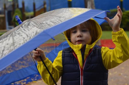 Foto de Niño lindo preescolar en un impermeable amarillo con un paraguas. Un niño juega afuera bajo la lluvia. Concepto Paseos divertidos de otoño o primavera, mal tiempo, pronóstico del tiempo, buen humor en un día nublado - Imagen libre de derechos
