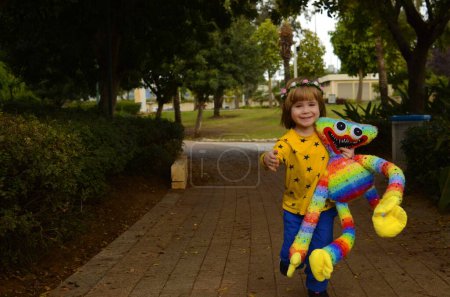 Foto de A cute boy in a yellow t-shirt runs along the path in the park with a large multi-colored soft toy. The child received a gift, the kid rejoices at a new friend. Concept: children and monsters, scary toys, imaginary friends. - Imagen libre de derechos