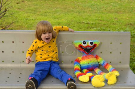 Foto de A positive little boy, 4 years old, walks in the park with a bright colored toy. Concept: kindergarten, imaginary friend, favorite toy. The child sits on a bench with a very large soft toy. - Imagen libre de derechos