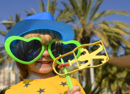 Téléchargez les photos : Carnaval, Pourim en Israël. Un petit garçon joyeux dans un chapeau, des lunettes drôles, une perruque avec un hochet, Raashan. Portrait d'un enfant en costume de Klana, des vacances drôles, un anniversaire, une mascarade. - en image libre de droit