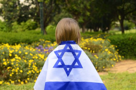 Little boy with the flag of Israel. A child in a blooming garden celebrates the Independence Day of the State of Israel. Child rear view, flag on the back.
