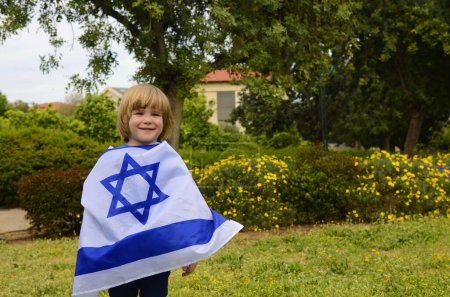 Little boy with the flag of Israel. A child in a blooming garden celebrates the Independence Day of the State of Israel. Child rear view, flag on the back.