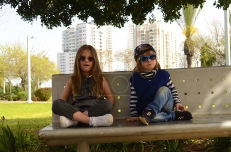 Téléchargez les photos : Des gosses cool. Un garçon et une fille sont assis sur un banc, les enfants dans des lunettes de soleil cachés à l'ombre. Frère et sœur, meilleurs amis. - en image libre de droit