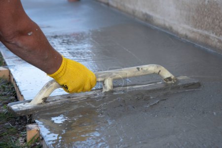 Mason leveling and screeding concrete floor base with square trowel in front of the house. Construction business, do-it-yourself concept. Hand in yellow glove.