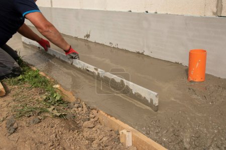 Mason leveling and screeding concrete floor base with square trowel in front of the house. Construction business, do-it-yourself concept. Hand in red glove.