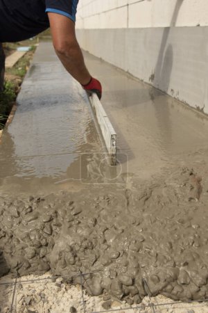 Mason leveling and screeding concrete floor base with square trowel in front of the house. Construction business, do-it-yourself concept. Hand in red glove