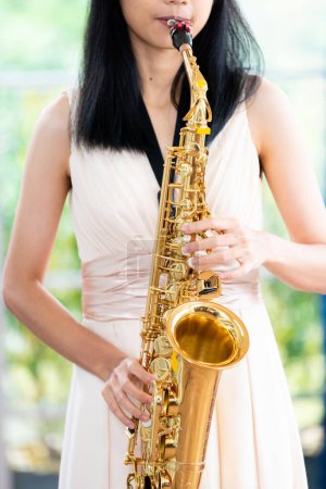 Jeune femme saxophoniste avec un instrument se tient près de la fenêtre. Intérieur confortable de la maison. Concept de passe-temps, monde intérieur, développement des talents, école de musique.