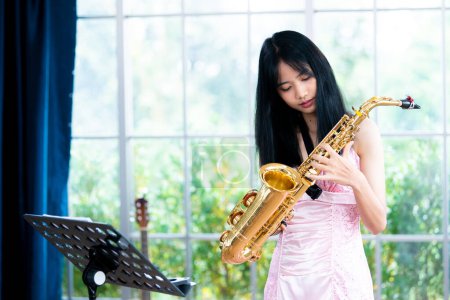 Jeune femme saxophoniste avec un instrument se tient près de la fenêtre. Intérieur confortable de la maison. Concept de passe-temps, monde intérieur, développement des talents, école de musique.