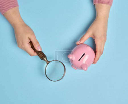 Photo for A woman's hand holds a piggy bank and a magnifying glass on a blue background, top view - Royalty Free Image