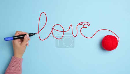 Une main de femme a écrit l'amour avec un feutre rouge et une boule de laine sur un fond bleu.
