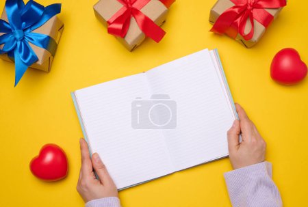Photo for An open notebook with empty white sheets and gift boxes around on a yellow background, top view. Festive background for wish - Royalty Free Image