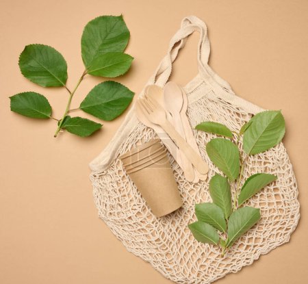 Téléchargez les photos : Sac à ficelle tricoté textile et une pile de verres en carton jetables en papier brun sur fond beige et feuilles vertes, vue de dessus - en image libre de droit