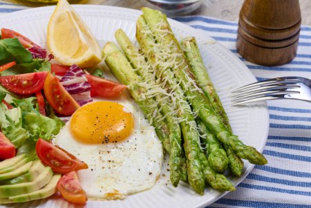 Foto de Placa redonda con espárragos cocidos, huevo frito, aguacate y ensalada de verduras frescas en la mesa, vista superior - Imagen libre de derechos