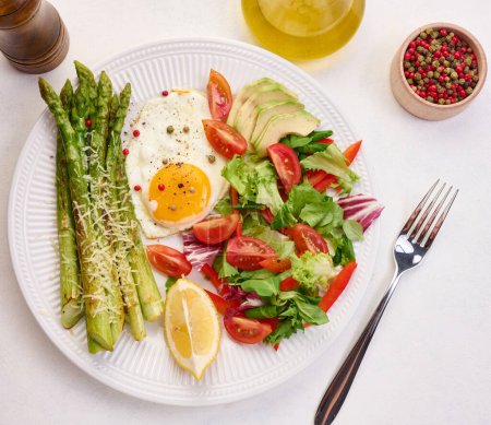 Foto de Placa redonda con espárragos cocidos, huevo frito, aguacate y ensalada de verduras frescas en la mesa, vista superior - Imagen libre de derechos