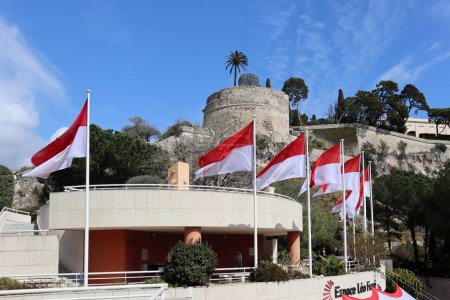 Foto de Mónaco, Mónaco - 19.03.2023: Banderas de Mónaco ondeando en el viento cerca de la roca con el castillo de Grimaldi - Imagen libre de derechos